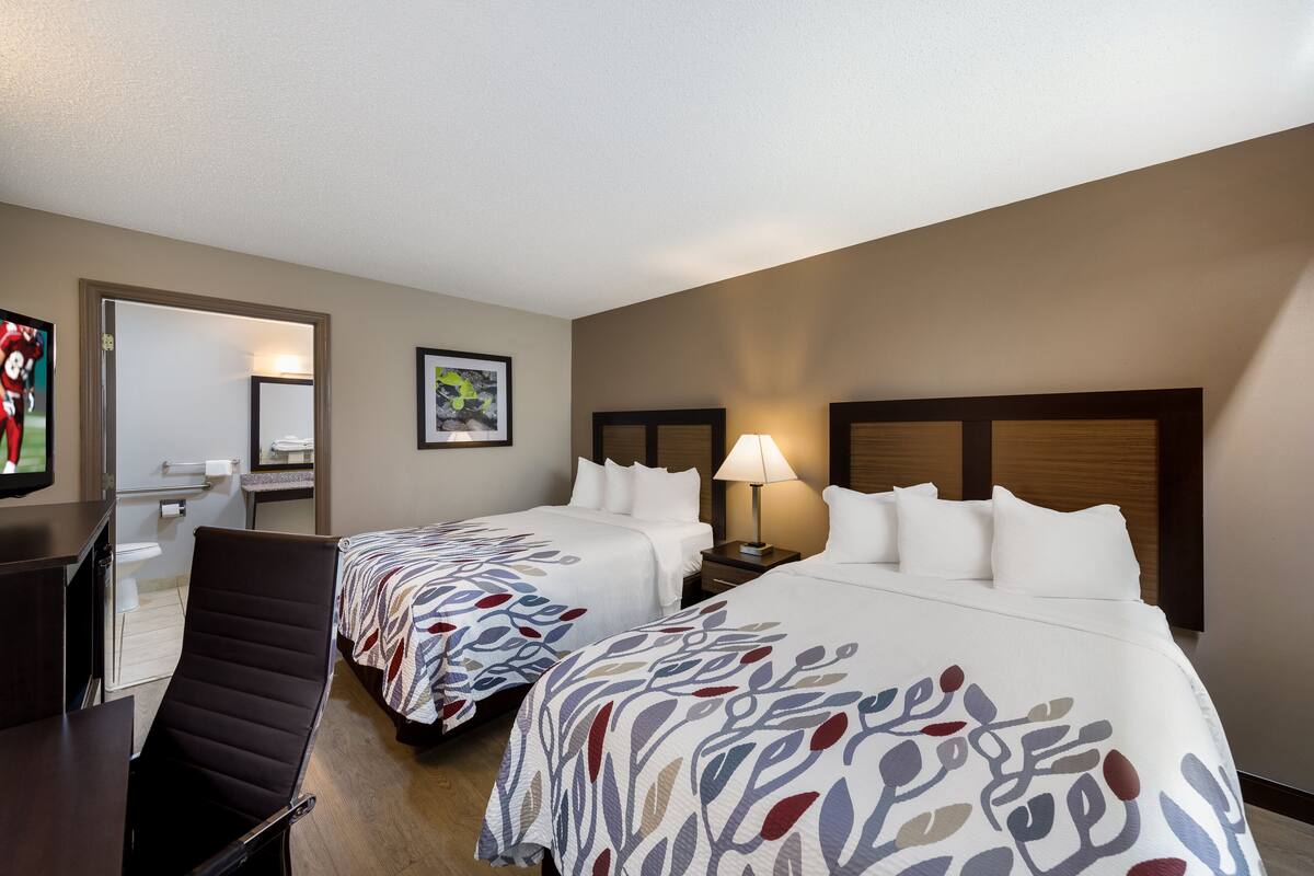 Quarto de hotel com duas camas, uma escrivaninha, uma cadeira, uma televisão e banheiro visível por uma porta aberta.