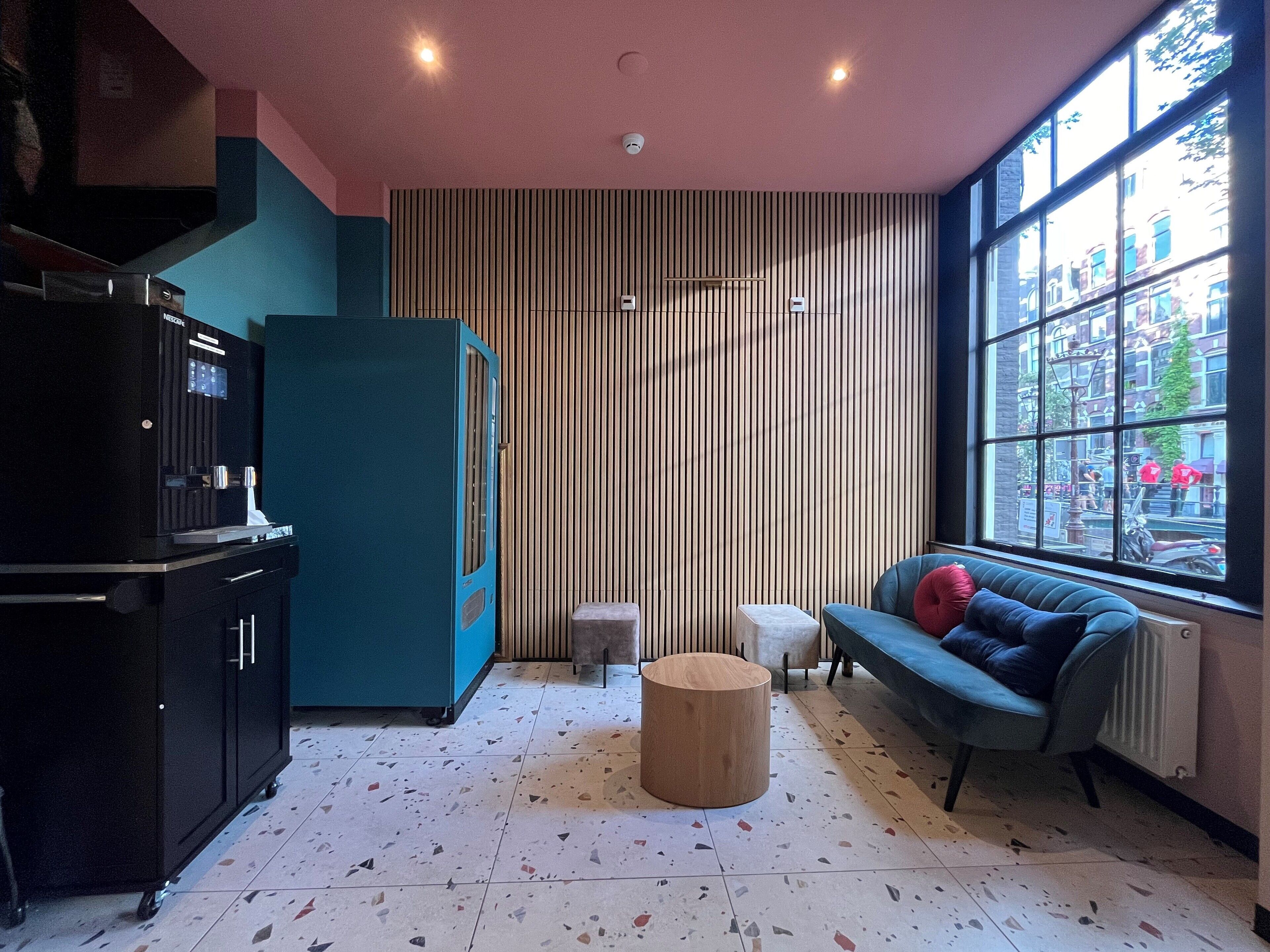 A modern living room with a sofa, a coffee table, a refrigerator, and a large window offering a view of the street.