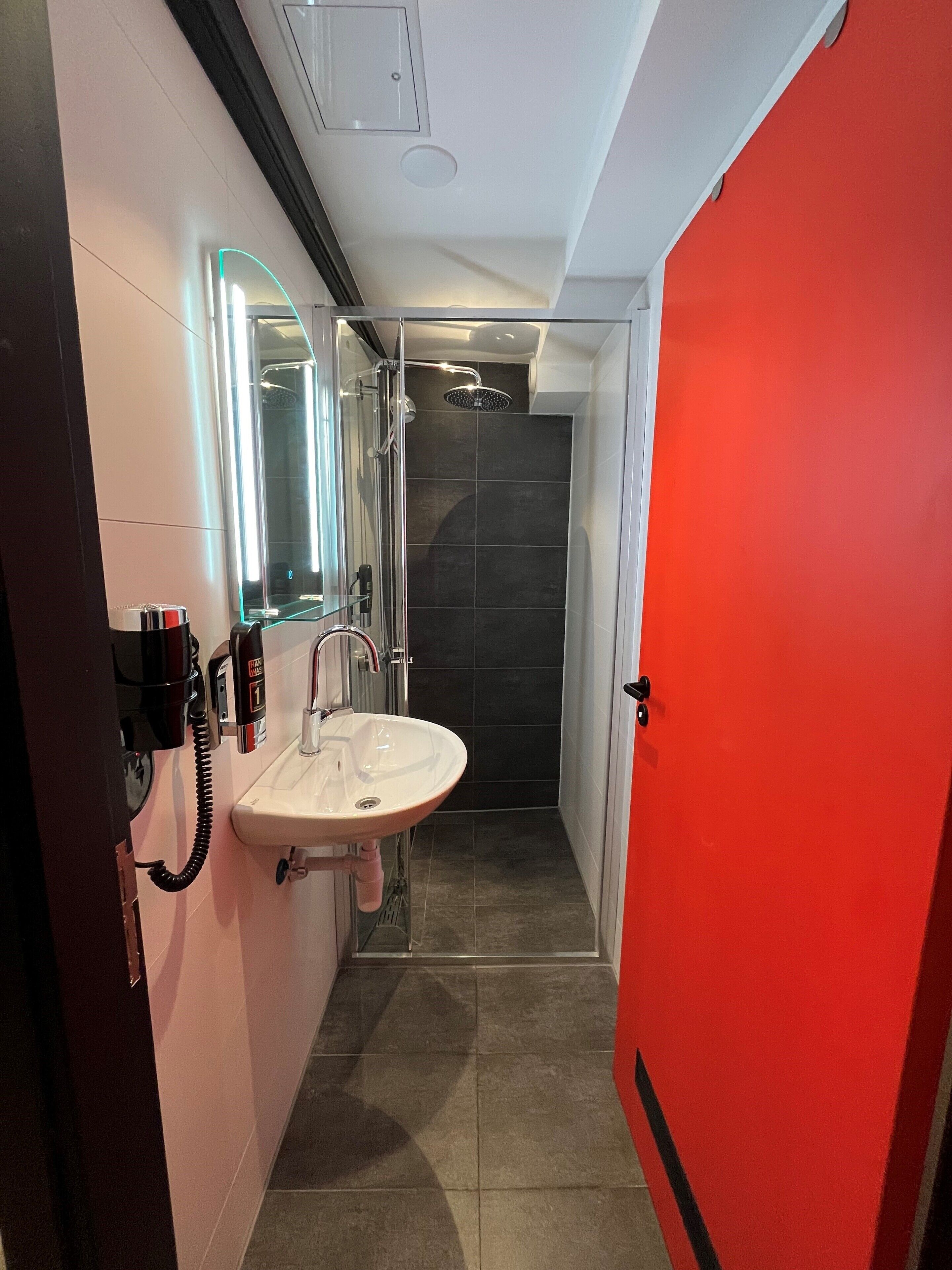A modern bathroom with a glass shower enclosure, a white sink, and a red door.