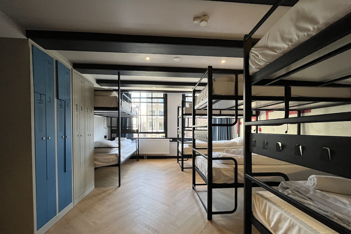 A dormitory room with bunk beds, a radiator, and a window with a view of buildings outside.
