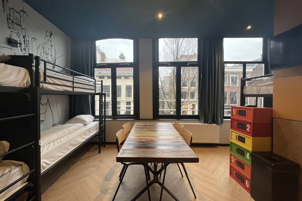A dormitory room with bunk beds, a wooden table, and a view of buildings through large windows.