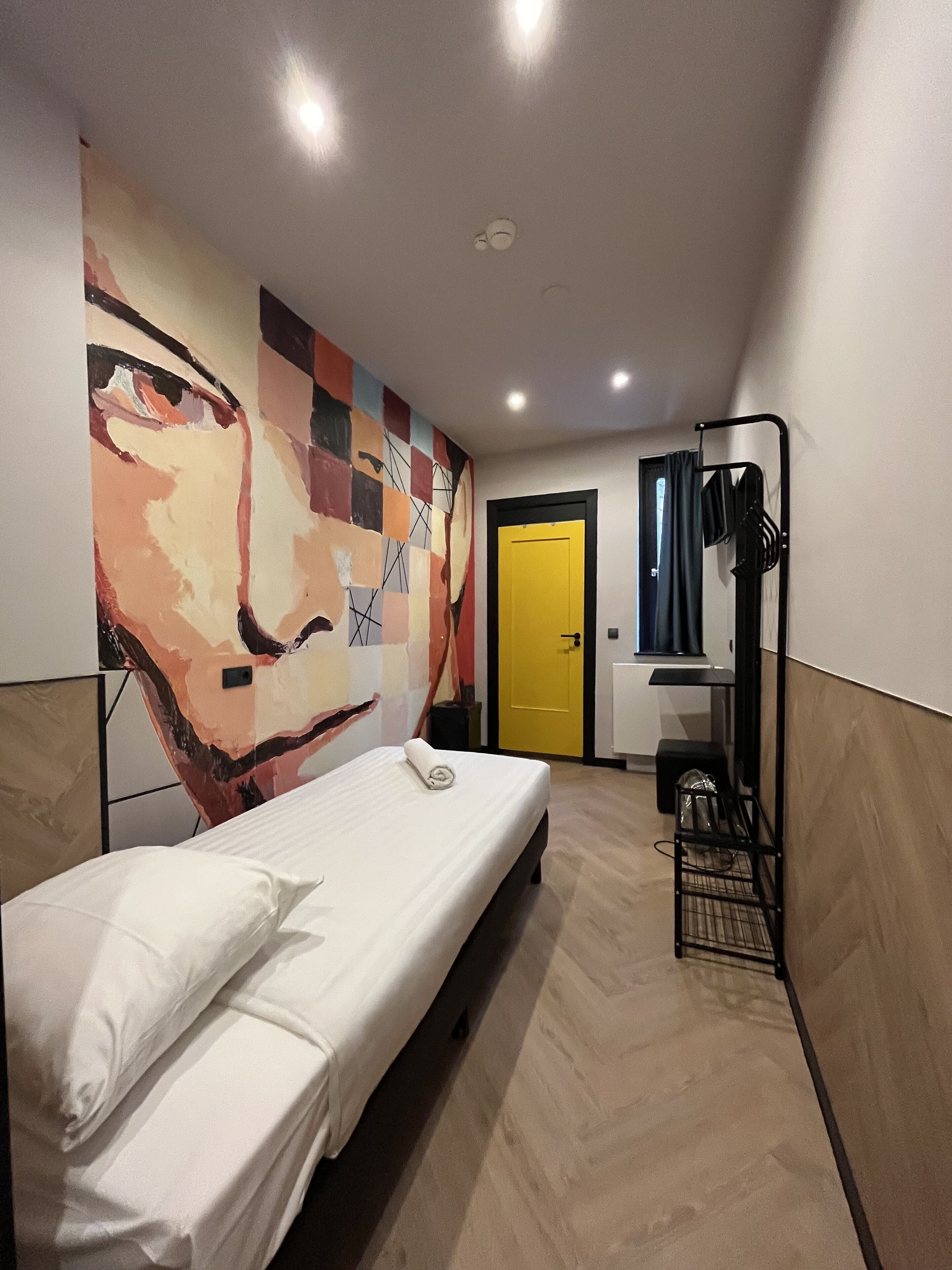 A single-bed room with a large abstract painting on the wall, a yellow door, and a metal rack with clothes.