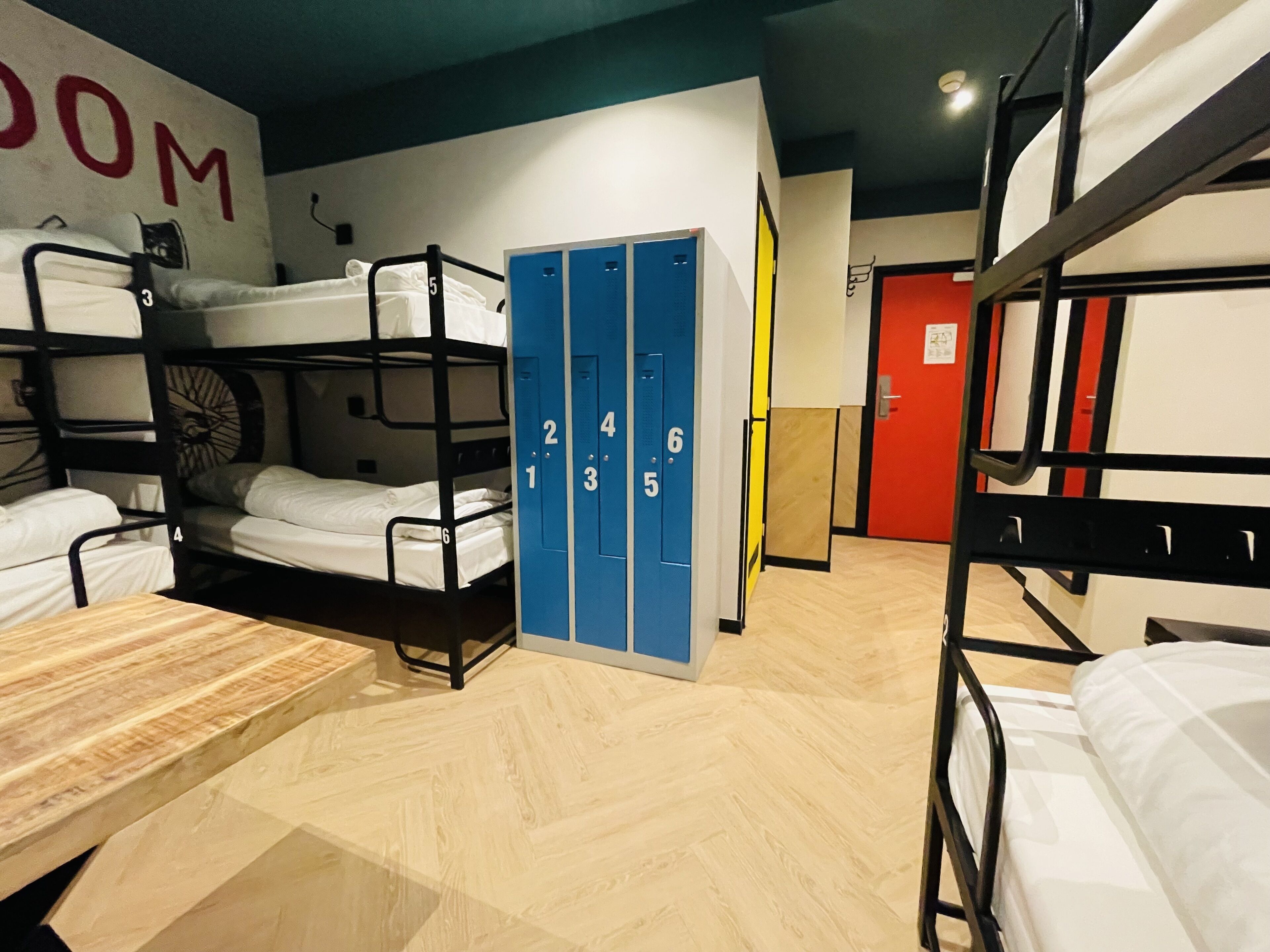 A dormitory room with bunk beds, a wooden table, and blue lockers numbered 1-6.