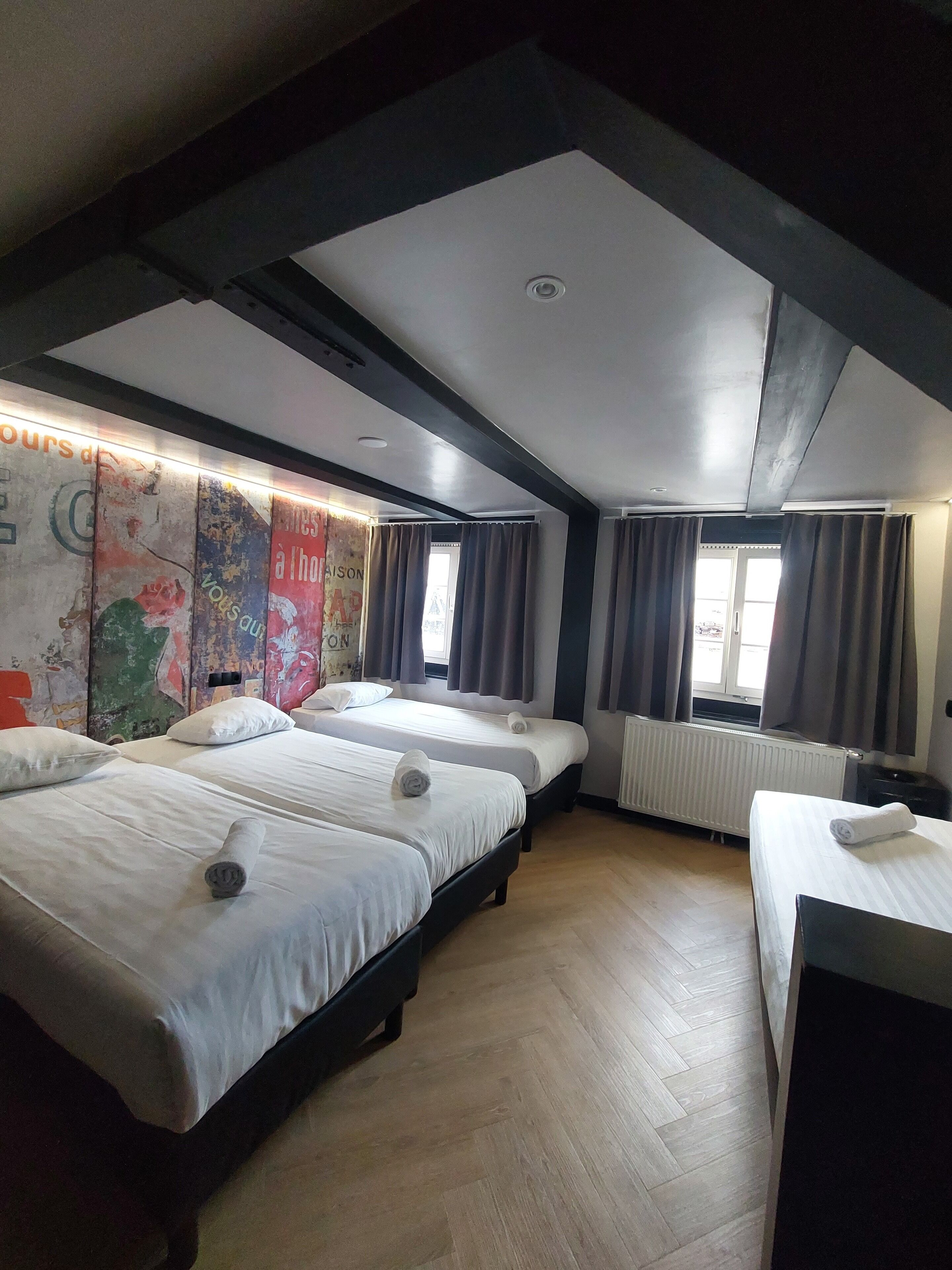Ein Hotelzimmer mit drei Betten, Holzboden, einem Heizkörper und einem Fenster mit Vorhängen.