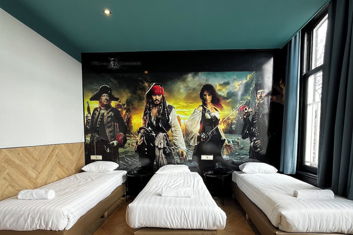 A hotel room with two single beds, a large poster of a pirate movie on the wall, wooden flooring, and a window with curtains.