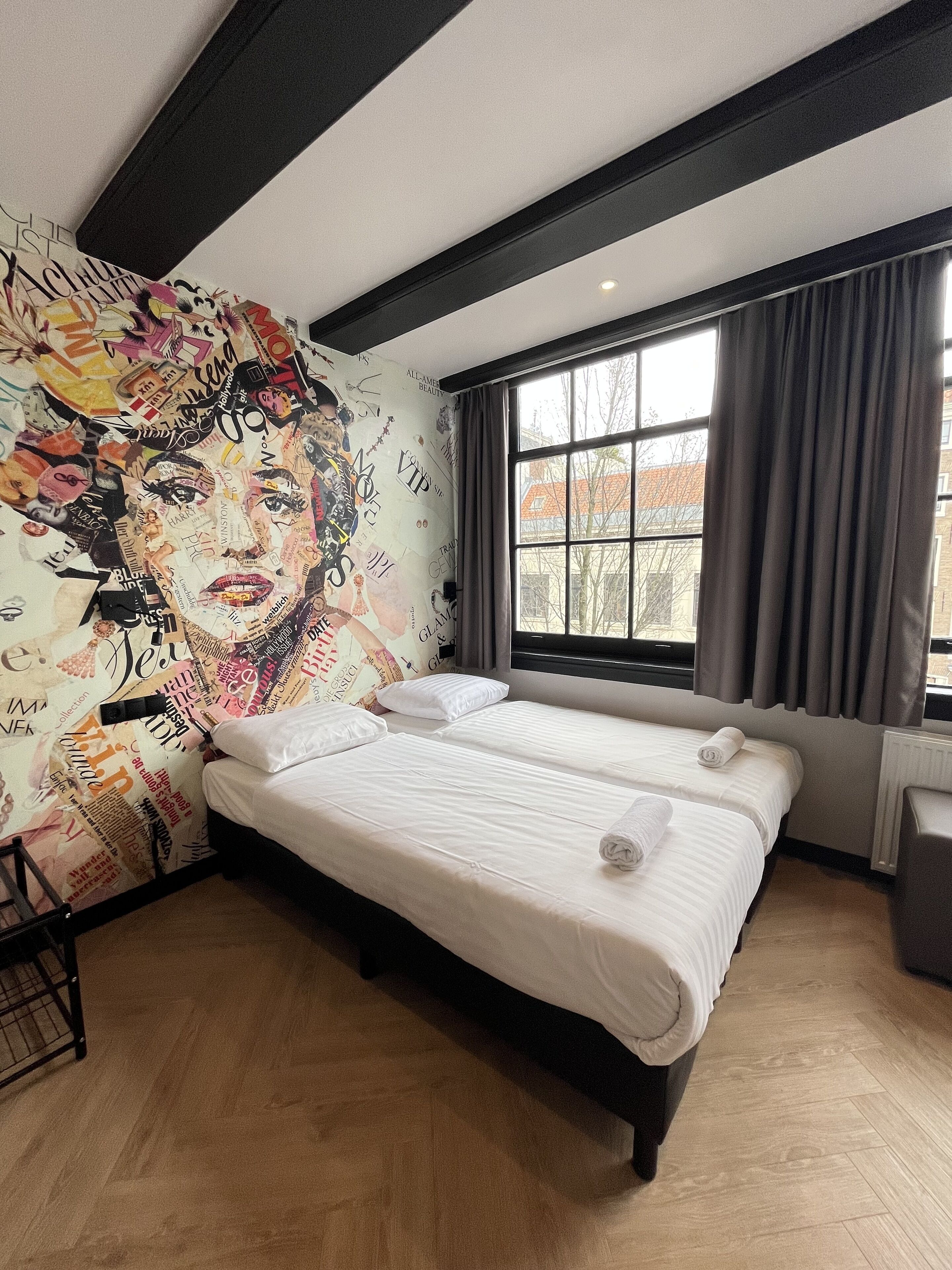 Habitación de hotel con dos camas, un mural grande en la pared, una ventana con cortinas y un radiador.