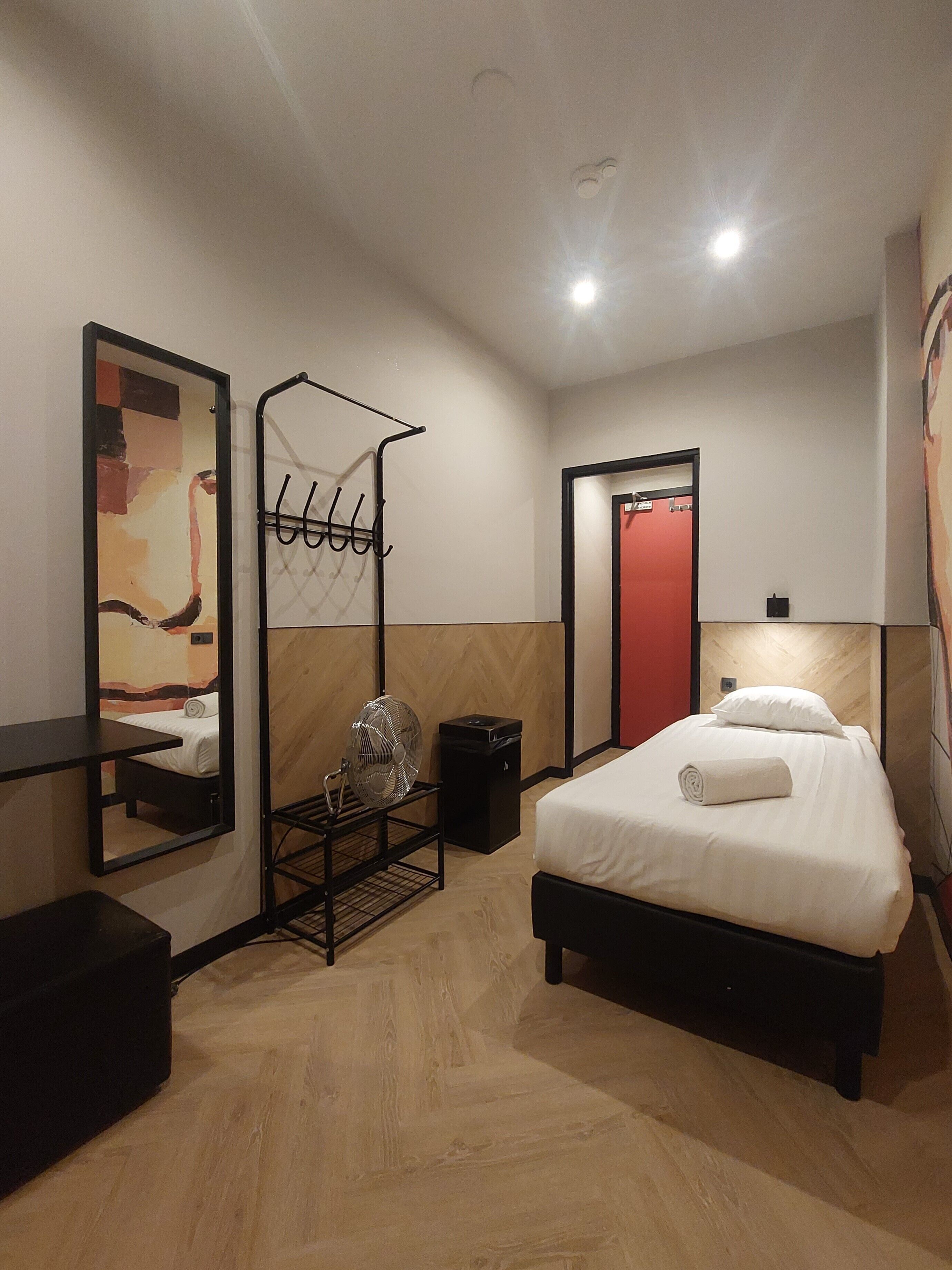 A hotel room with a bed, a bedside table, a mirror, a fan, and a red door.