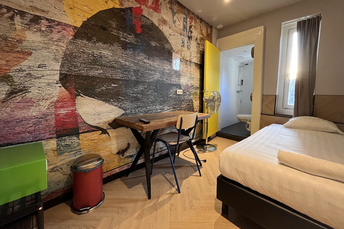 A hotel room with a bed, a desk, a chair, a trash bin, and a bathroom visible through an open door.