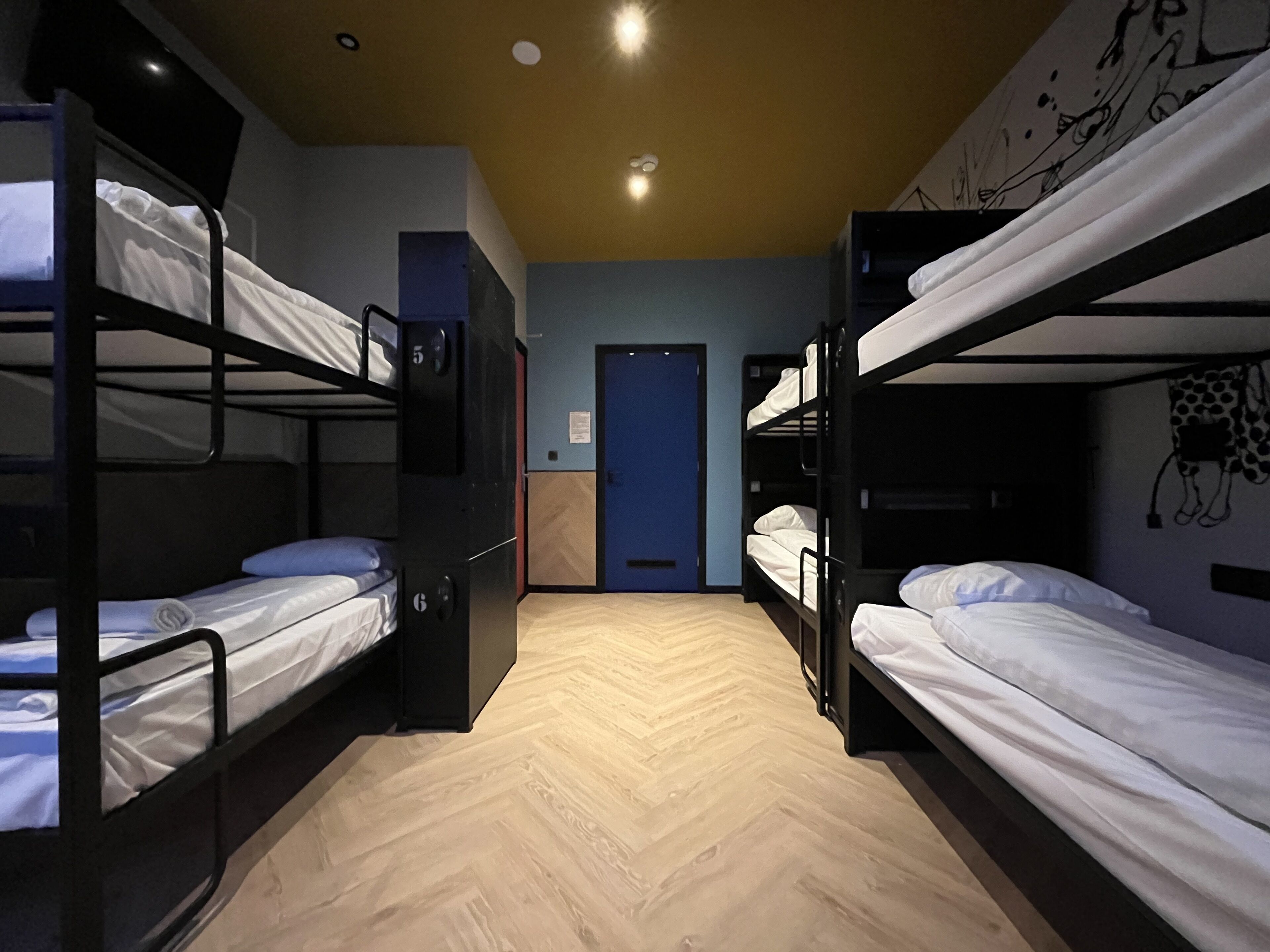 Una habitación con literas, piso de madera tipo espiga, una puerta azul y un mural de jirafa en la pared.