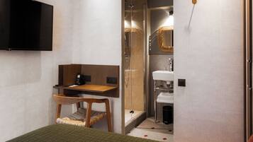 Habitación de hotel compacta con cama, escritorio, silla, televisor montado en la pared, zona de ducha y baño con espejo y lavabo.