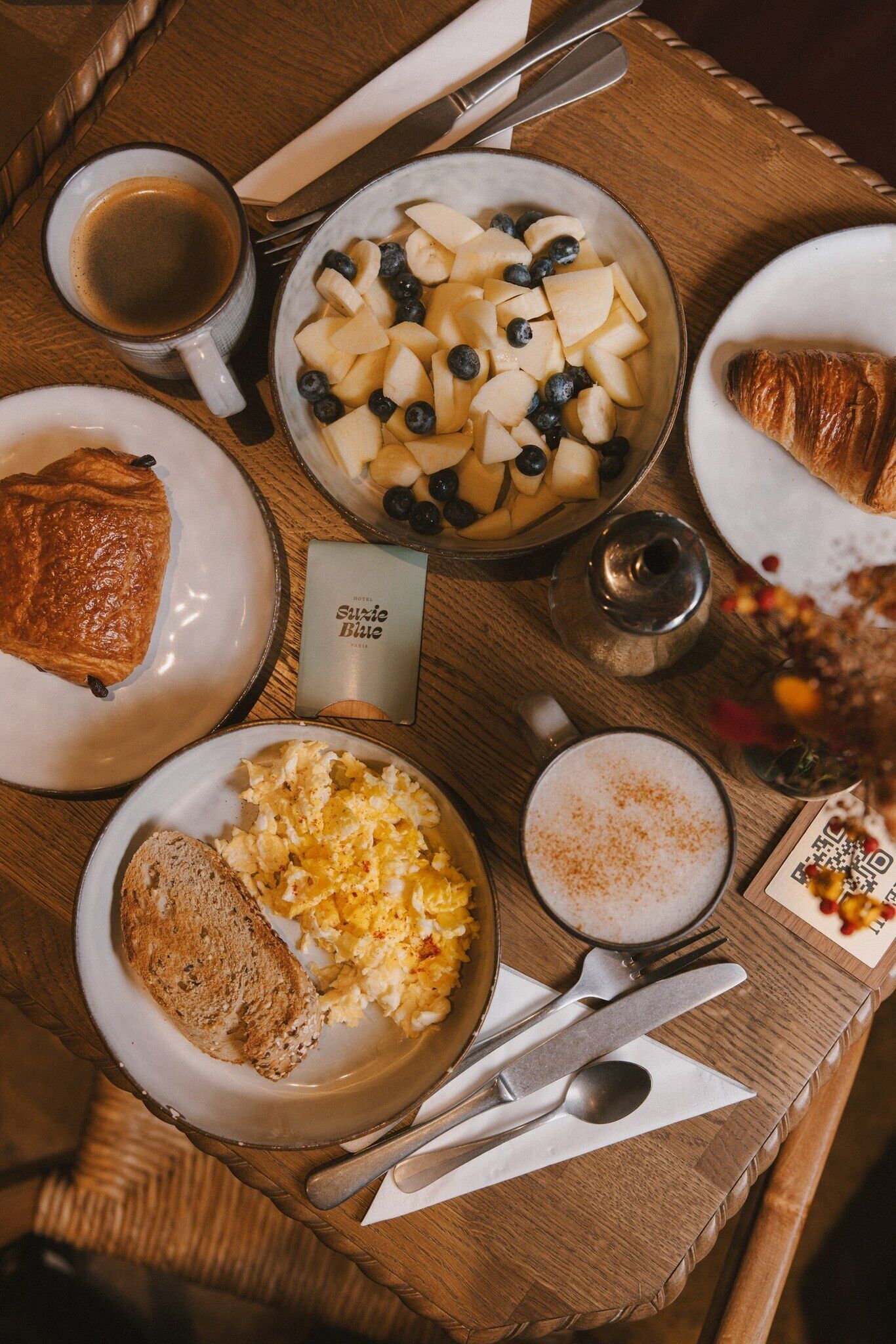 A breakfast tray with various food items including toast, scrambled eggs, a fruit bowl with apples and blueberries, a croissant, and a cup of coffee.