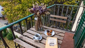Un banc en bois, sur lequel se trouve un vase de fleurs séchées, une assiette de muffins, une tasse de café et un livre intitulé « La strada di Swann », sur un balcon.