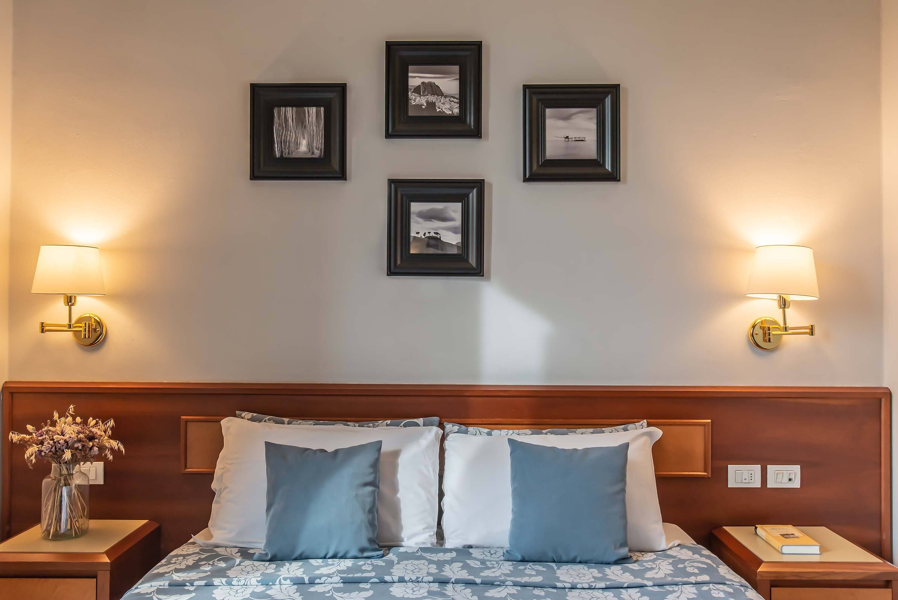 A hotel room with a wooden headboard, a bed with blue and white bedding, two bedside tables with lamps, and four framed pictures on the wall.