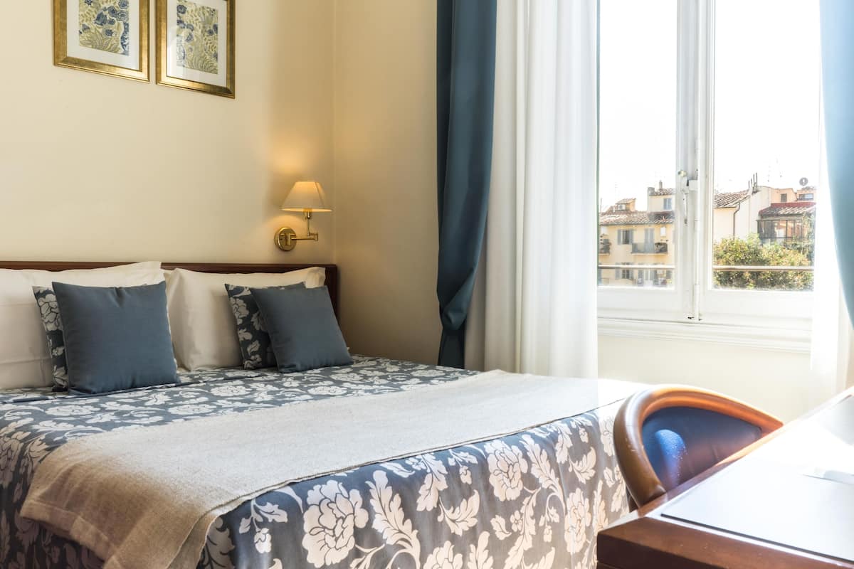 Habitación de hotel con cama, escritorio, lámpara y ventana con vistas a edificios.