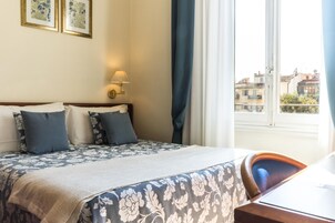 A hotel room with a bed, a desk, a lamp, and a window with a view of buildings.