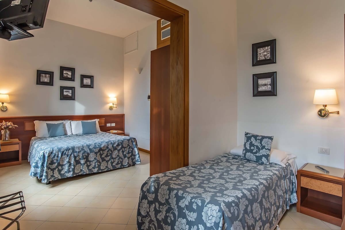 Habitación de hotel con dos camas, cabecera de madera, mesita de noche, lámpara y cuadros en la pared.