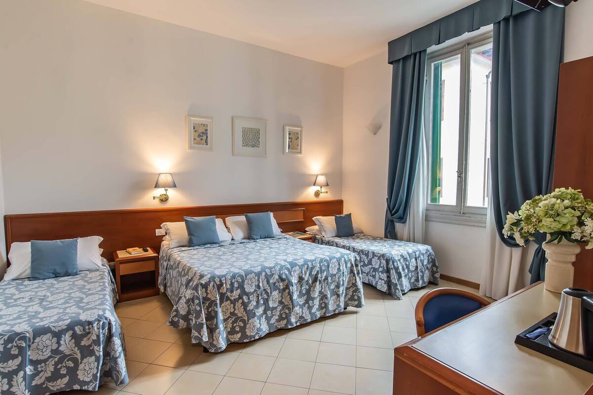 Habitación de hotel con dos camas, un escritorio y una silla. Hay lámparas de pared y una ventana con cortinas.