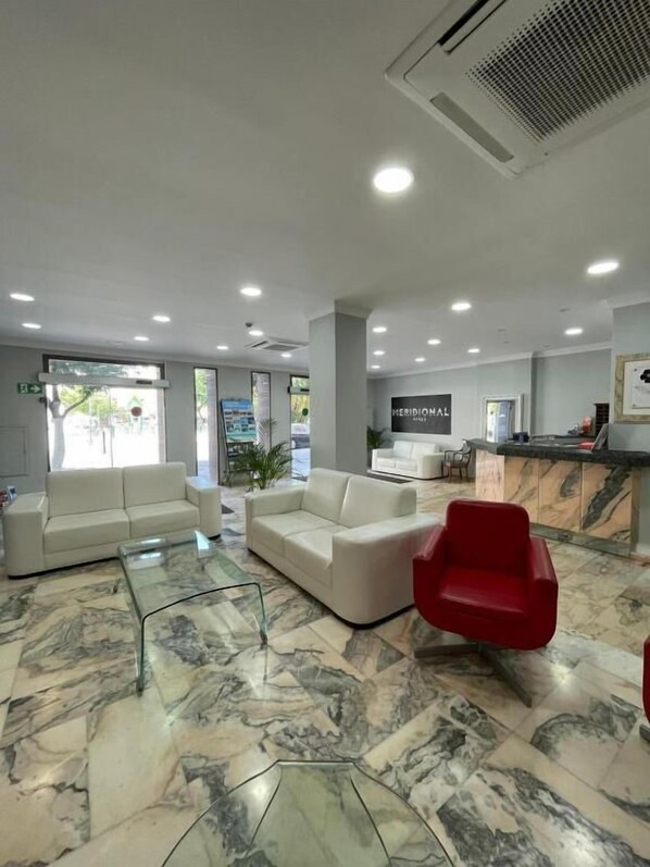 Un hall d’hôtel avec un sol en marbre, deux canapés blancs, un fauteuil rouge et un comptoir d’accueil.