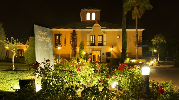 Un hotel con una entrada destacada, un balcón y un jardín bien iluminado con flores rojas.