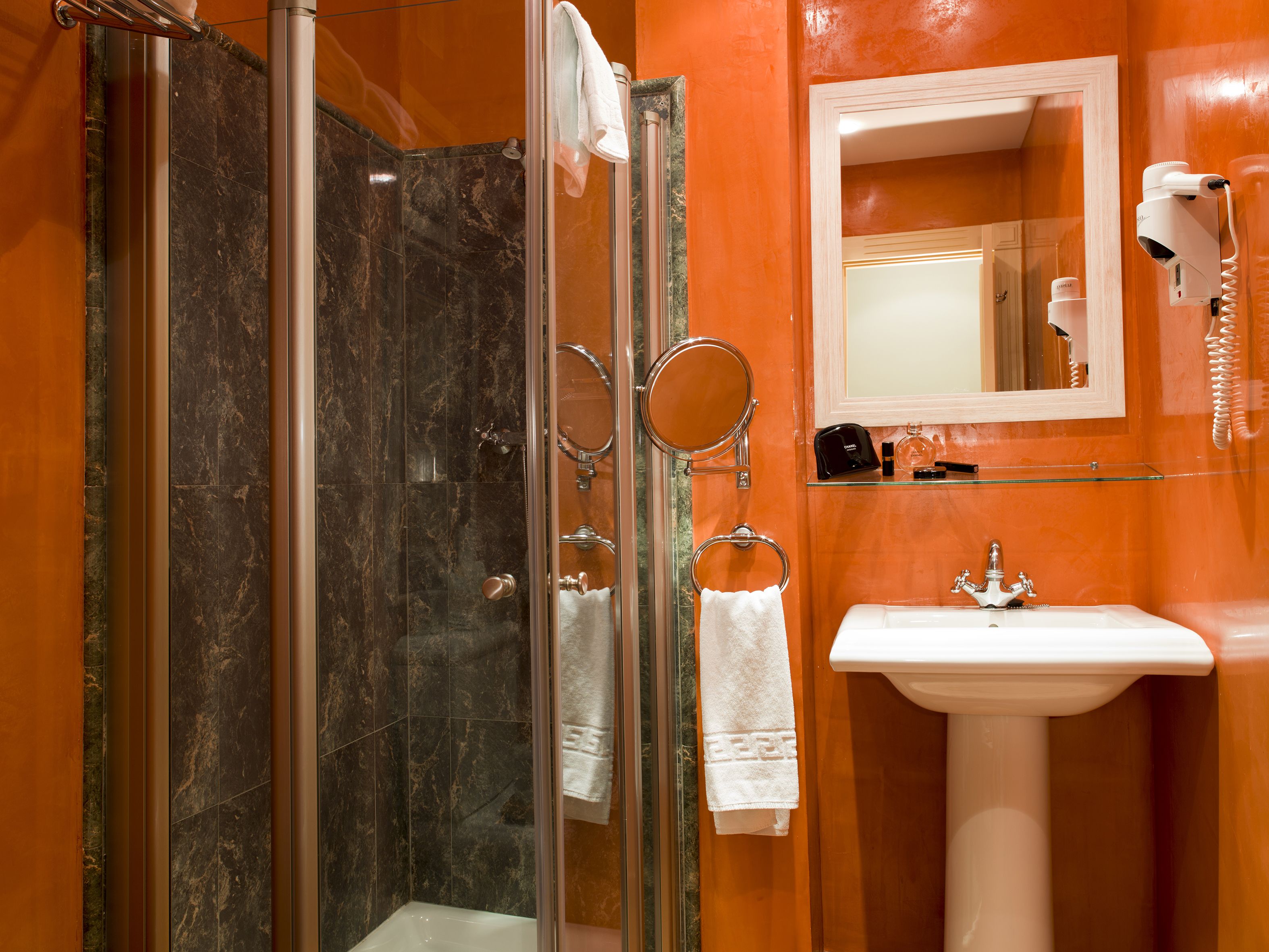 A bathroom with a glass shower enclosure, a sink, a mirror, and a hairdryer on the wall.