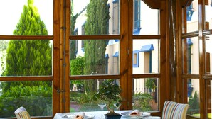 Un comedor con una mesa lista para dos, ventanas amplias y vistas a edificios y árboles.
