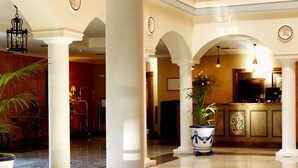 Un hall de hotel con columnas blancas, una recepción, una zona de bar y un comedor con pared roja y una araña.