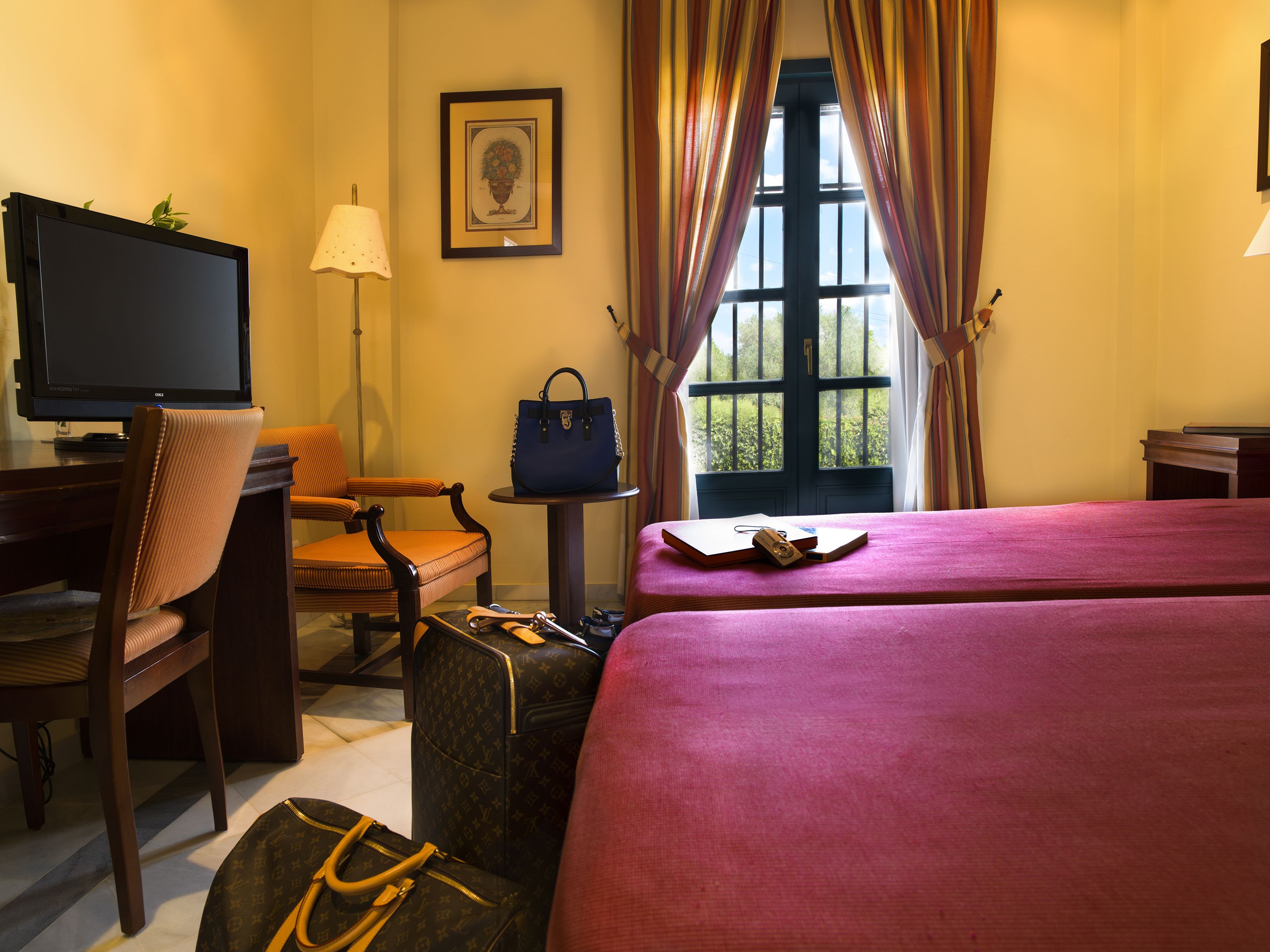 A hotel room with a bed, a chair, a desk with a television, a suitcase, and a window with curtains.