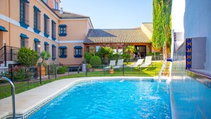 Una piscina con borde alicatado, rodeada por una valla, y contigua a un edificio con techo y marcos de ventana azules.
