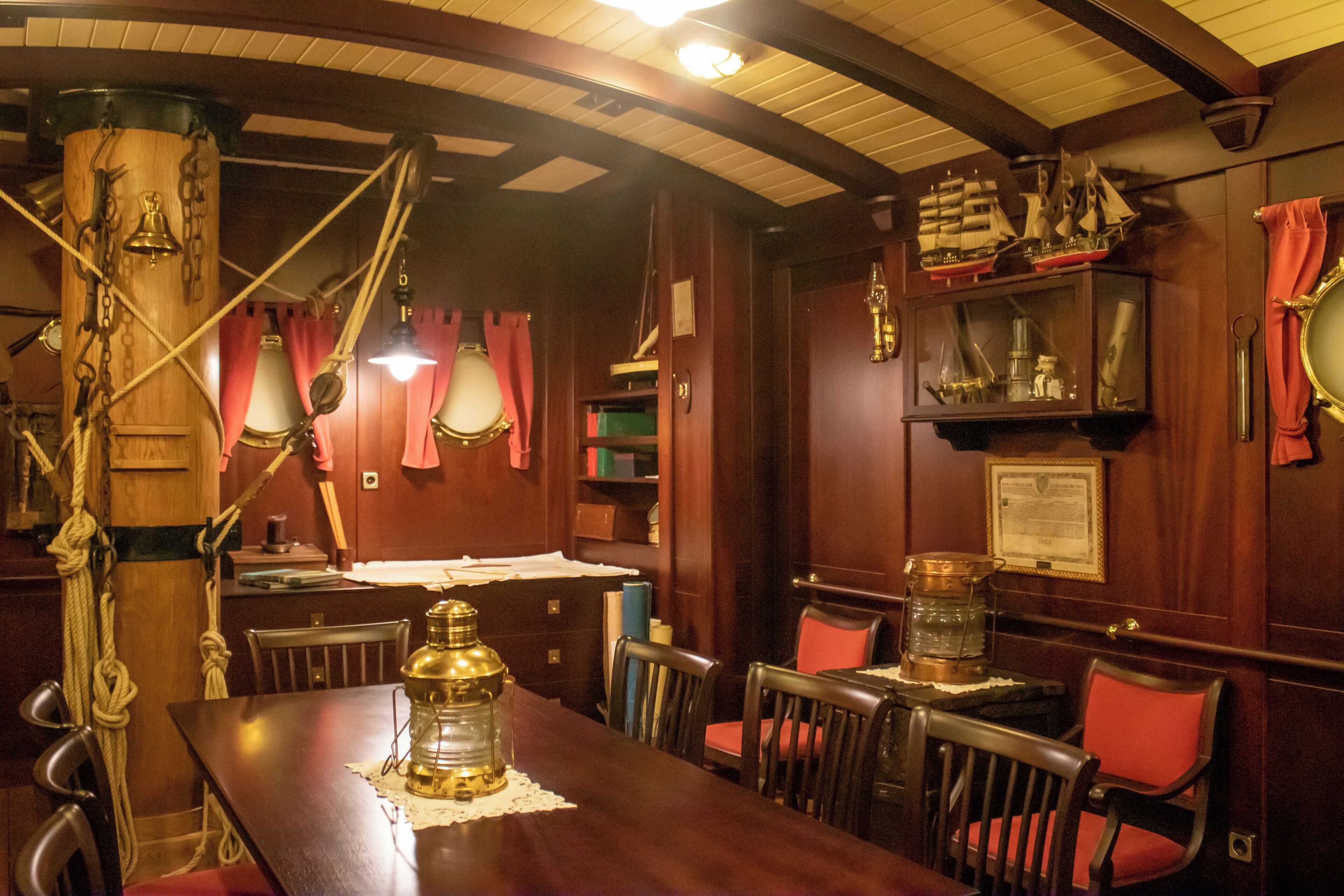 A wooden interior with a long table, chairs, and nautical-themed decor including a ship model and brass instruments.