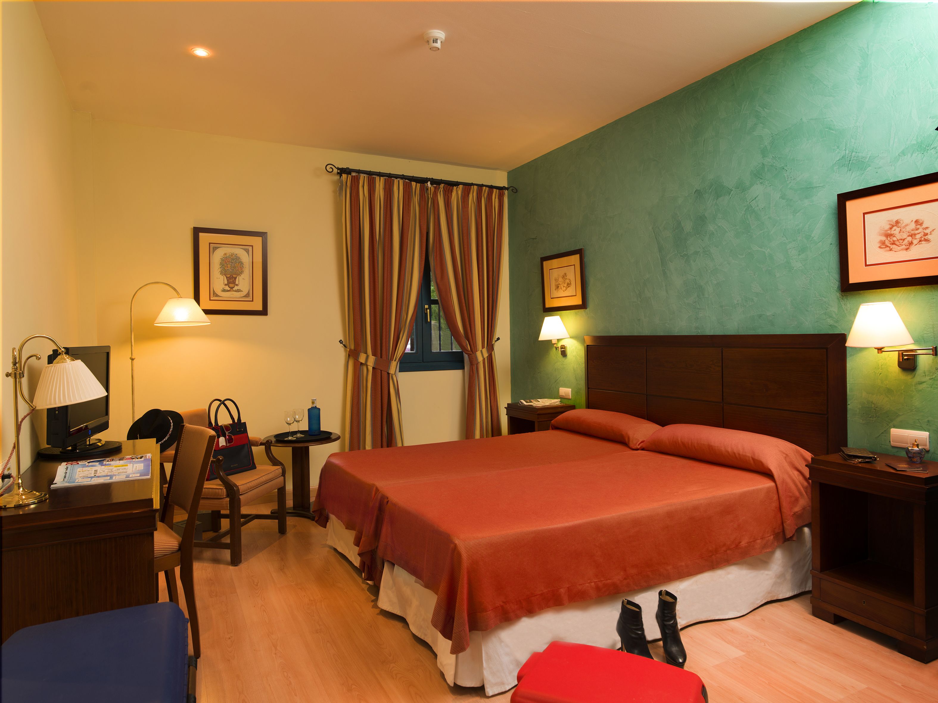 Habitación de hotel con una cama grande, un escritorio con computadora, una silla, una mesita con lámpara y una ventana con cortinas.