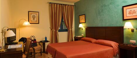 A hotel room with a large bed, a desk with a computer, a chair, a small table with a lamp, and a window with curtains.