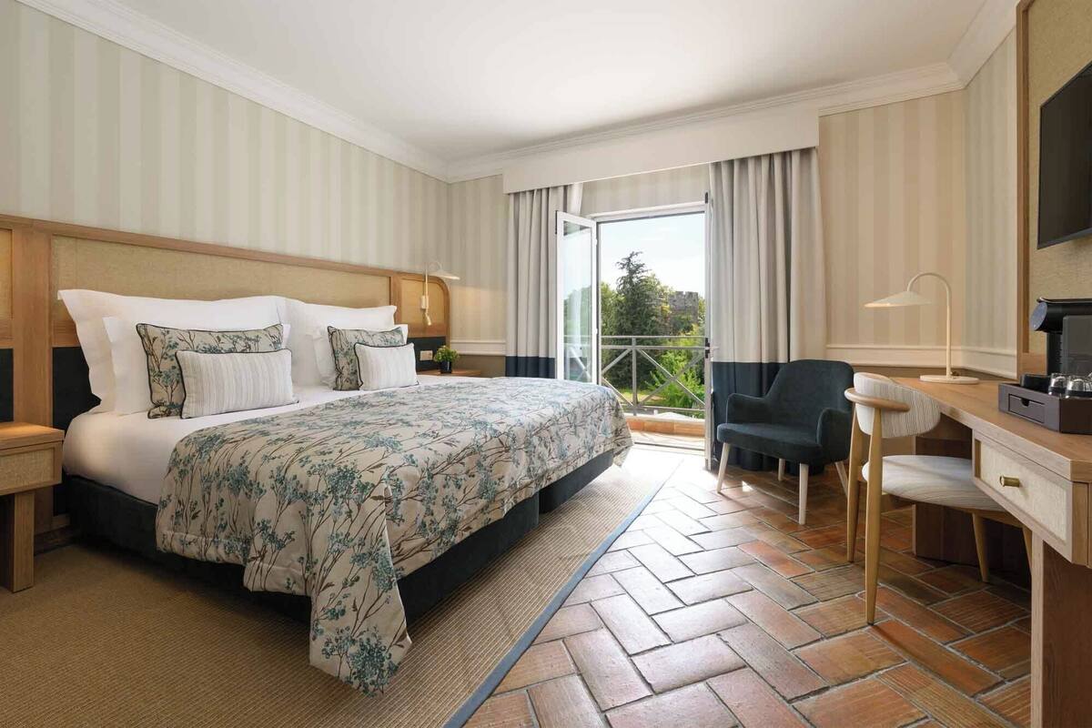 A hotel room with a large bed, a desk, a chair, and a balcony with curtains.