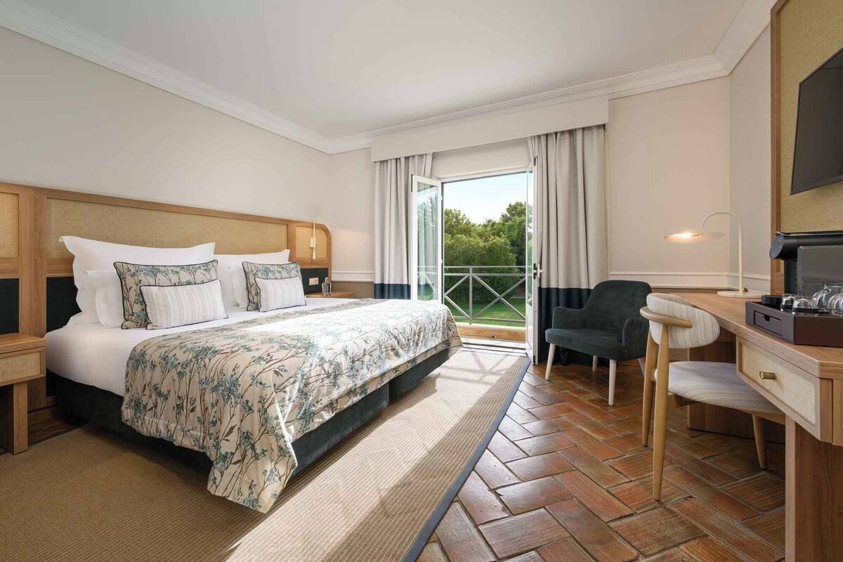 A hotel room with a large bed, a desk with a chair, a balcony with curtains, and a view of greenery.