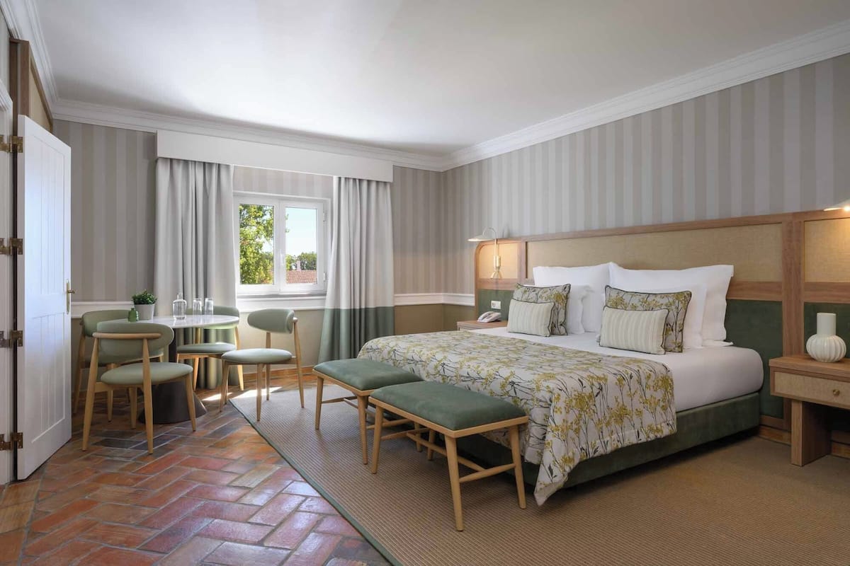 A hotel room with a large bed, bedside tables, a desk with chairs, a window with curtains, and a patterned floor.