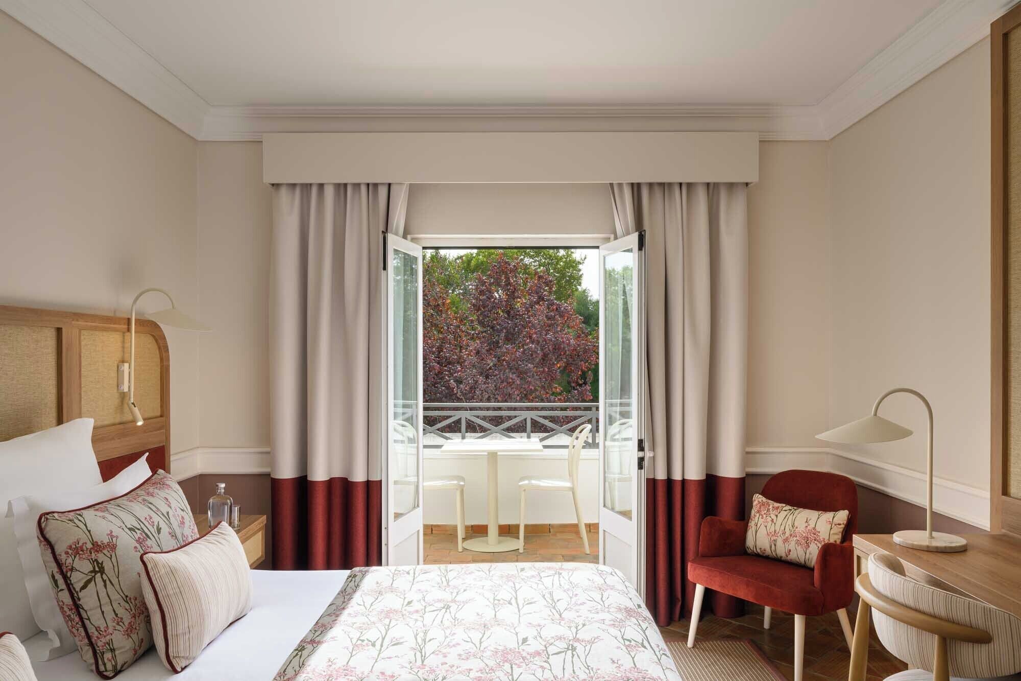 A hotel room with a bed, a desk, a chair, and a balcony with a view of trees.