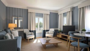 Una habitación de hotel moderna con sofá, dos sillones, una mesa de centro, televisor y un área de comedor con sillas y mesa.