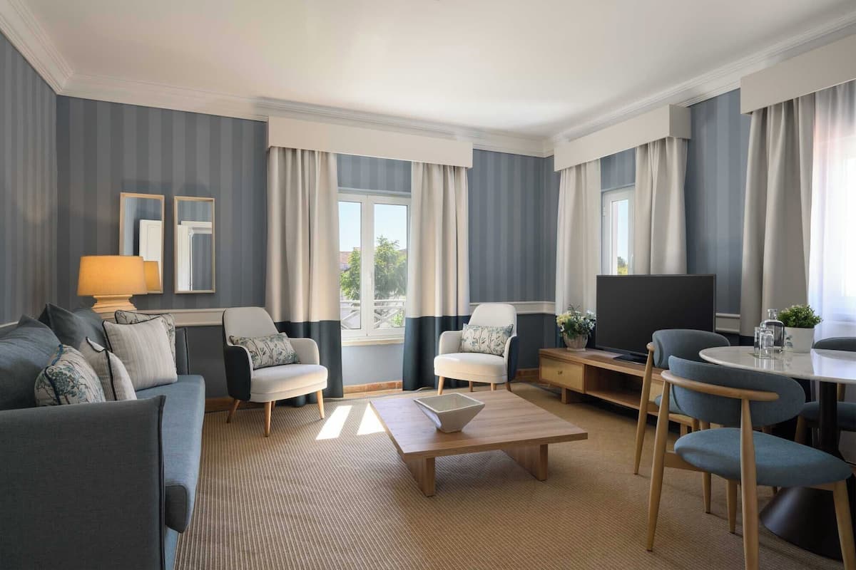 A modern hotel room with a sofa, two armchairs, a coffee table, a TV, and a dining area with chairs and a table.