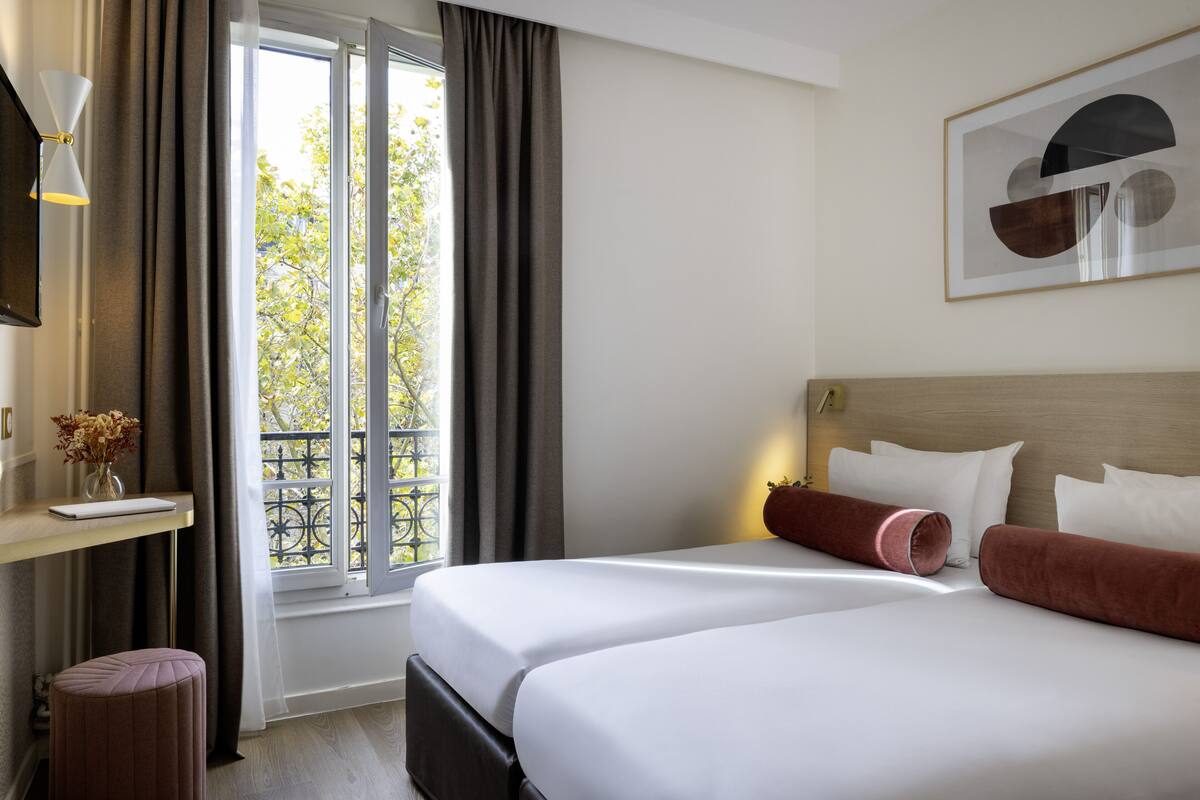Een hotelkamer met twee bedden, een bureau met lamp, een raam met gordijnen en een lijst met afbeelding aan de muur.
