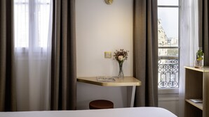 A hotel room with a bed, a desk with a lamp, a small stool, a vase with flowers, and a view of the city through a window. - Hotel Bailli de Suffren (Paris)
