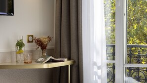A hotel room with a bed, a bedside table with a lamp, a small stool, a window with a view of trees, and a wall-mounted TV. - Hotel Bailli de Suffren (Paris)