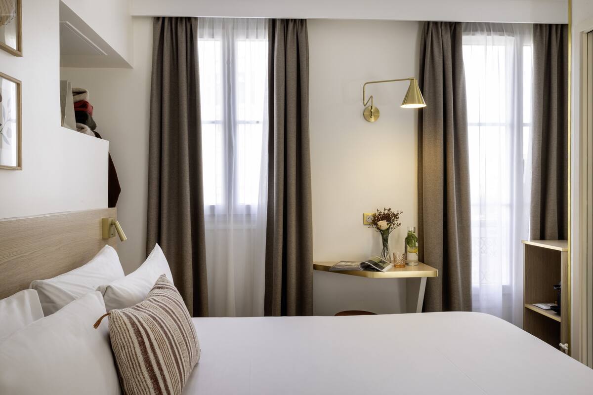 Een hotelkamer met een bed, nachtkastje met lamp, een klein bureau en een plank.
