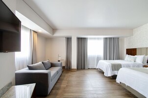 Una habitación de hotel moderna con sofá, dos camas, un televisor de pantalla plana y un ventanal con cortinas.