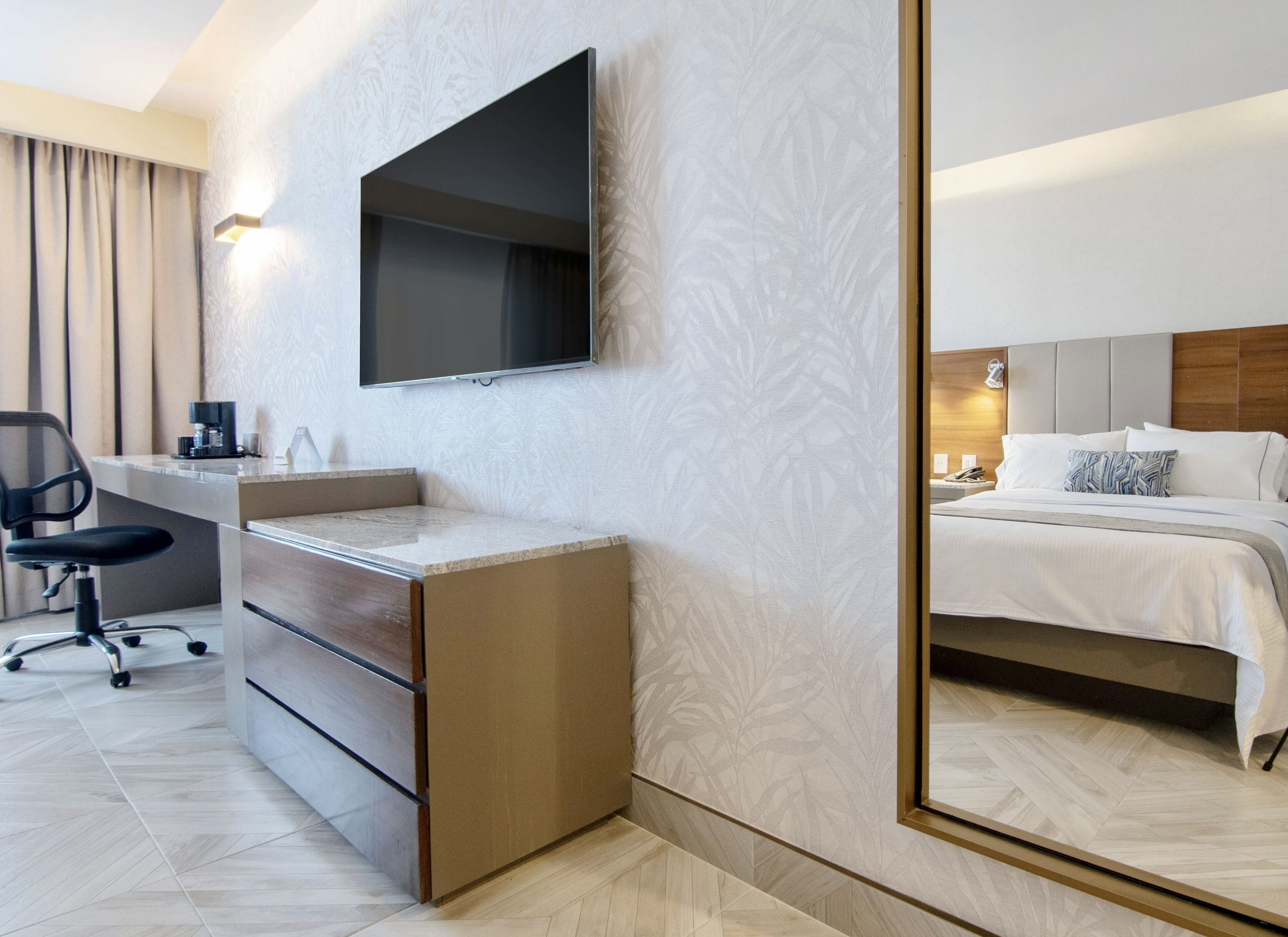 A hotel room with a flat-screen TV mounted on the wall, a desk with a chair, a bed with white bedding, and a large mirror reflecting the room.
