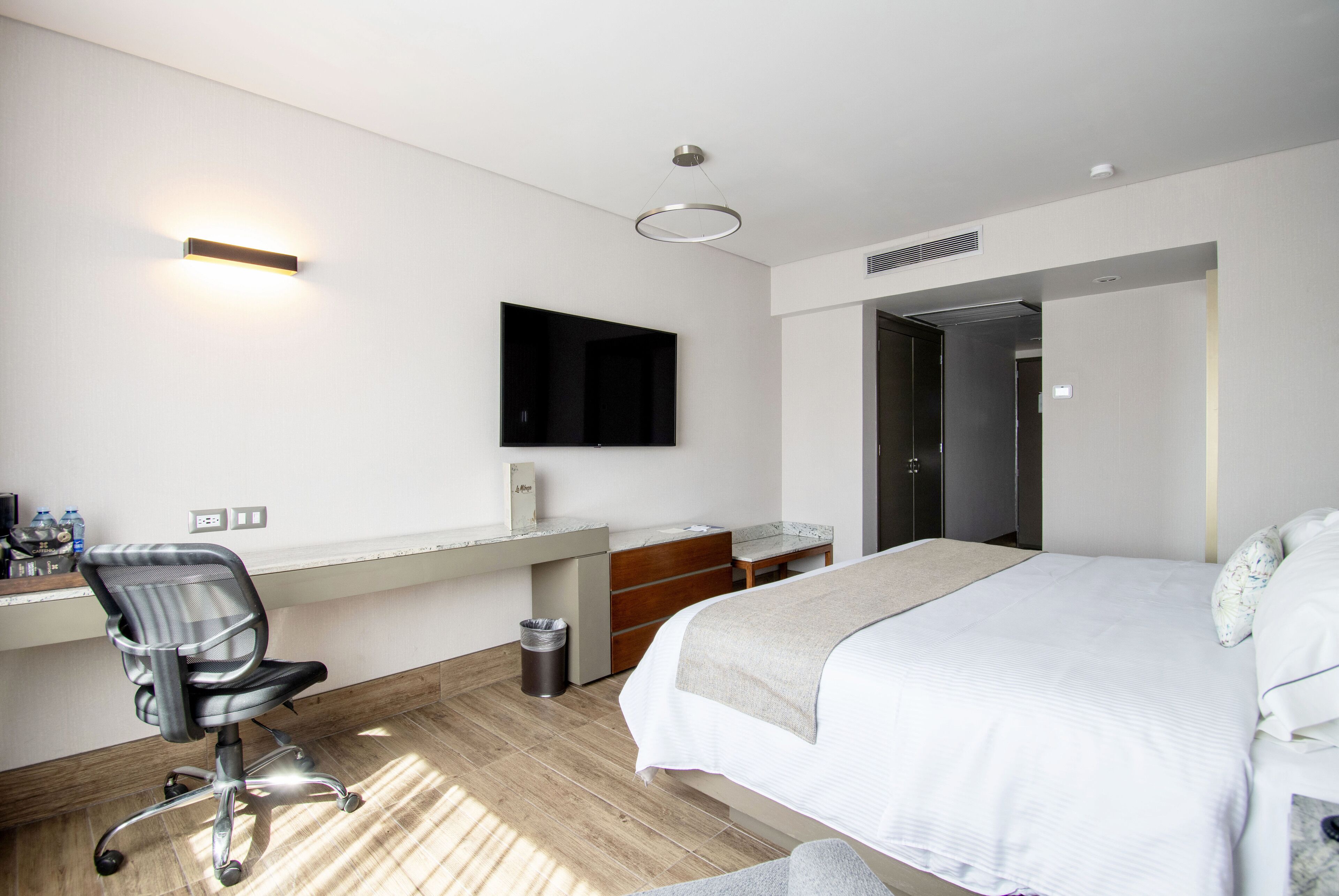 A modern hotel room with a bed, desk, chair, and a TV mounted on the wall.