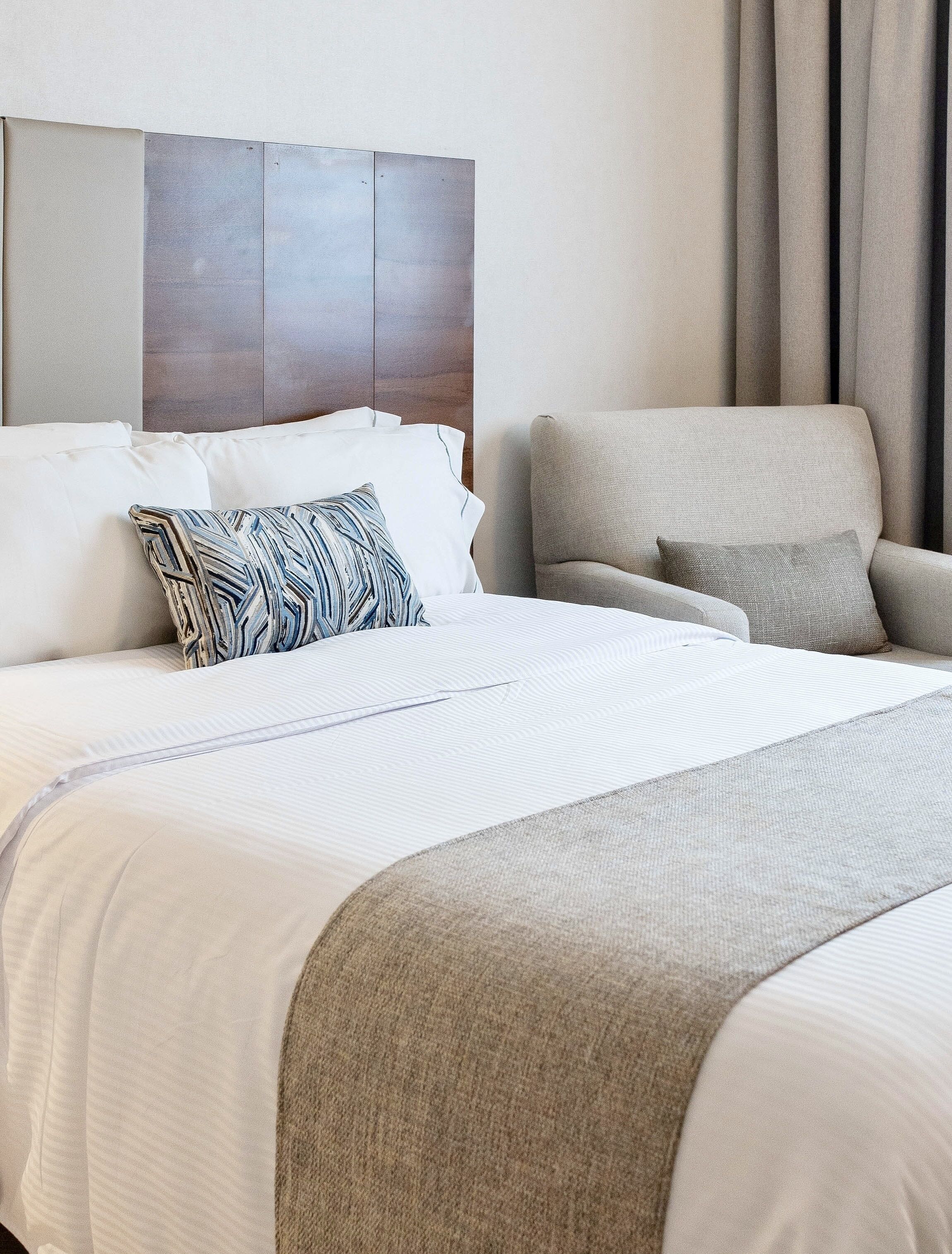 Quarto de hotel com uma cama grande, cabeceira de madeira, uma poltrona cinza e uma janela com cortinas.