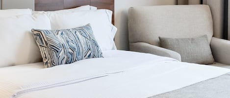 Habitación de hotel con una cama grande, cabecero de madera, un sillón gris y una ventana con cortinas.