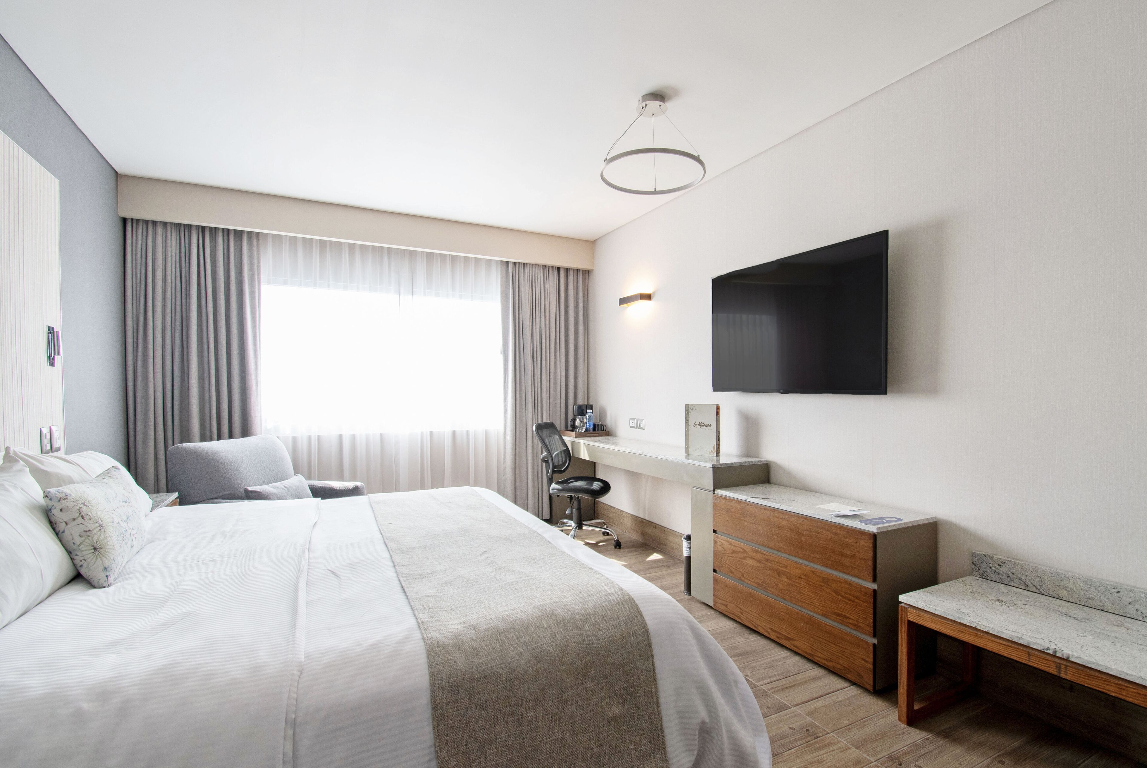 Une chambre d’hôtel moderne, dotée d’un grand lit, d’un bureau, d’une chaise, d’un téléviseur fixé au mur et d’une fenêtre avec des rideaux.