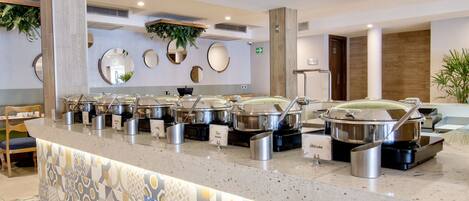 Zona de bufé con varias estaciones de autoservicio, que ofrecen opciones de comida caliente y fría, y un área de comedor con piso de baldosas estampadas.