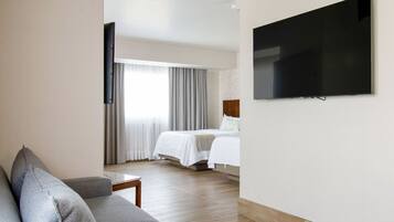 A modern hotel room with a grey sofa, a flat-screen TV mounted on the wall, a bed with white bedding, and a large window with curtains.