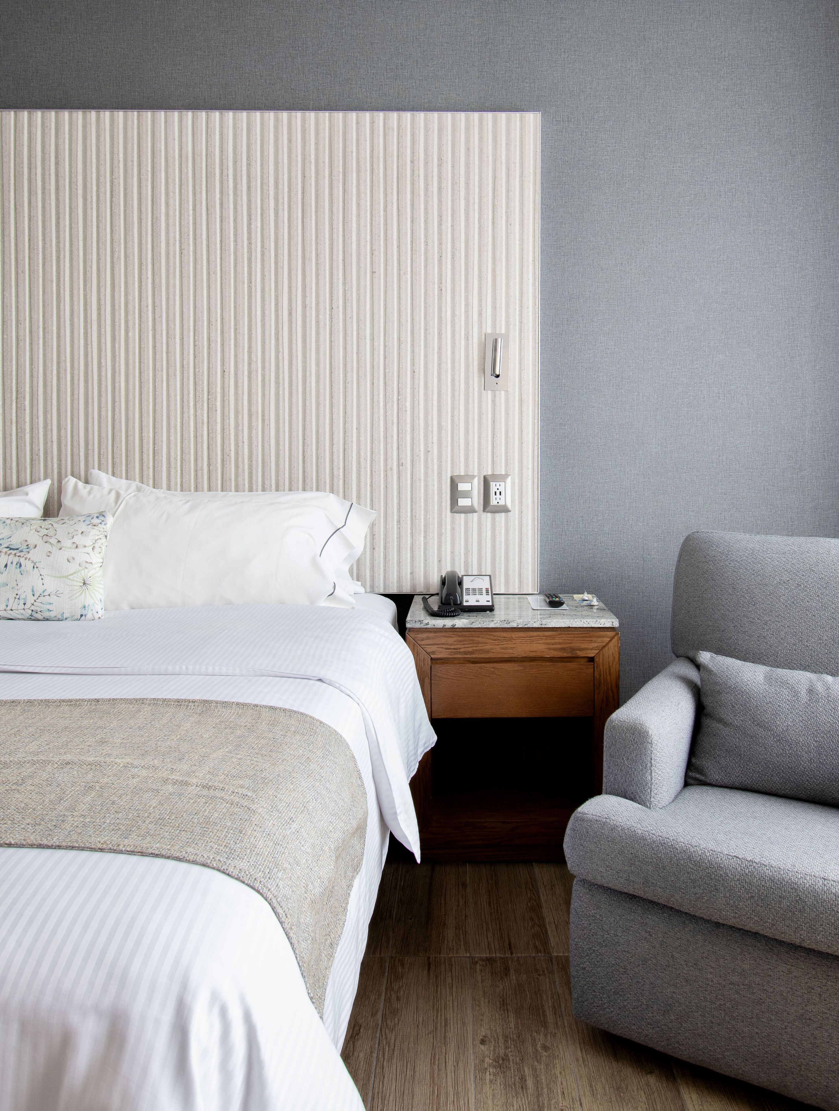 A hotel room with a large bed, a bedside table with a phone, and a grey armchair.